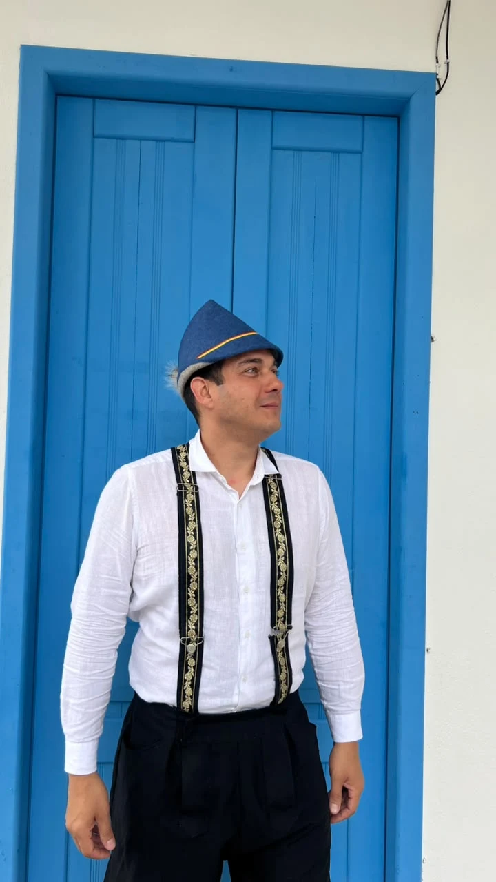 Hans Dettmann de perfil diante de uma porta azul, com camisa branca e chapéu azul.